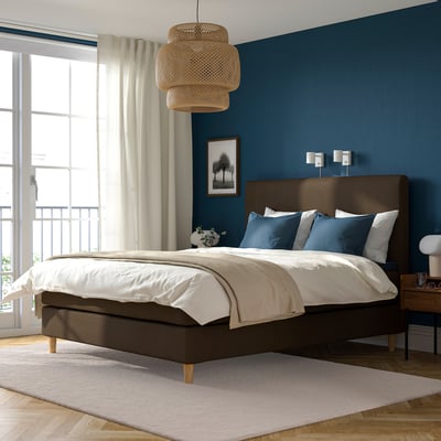 Modernes, dunkelbraunes Bett, groß, mit blauen Kissen, Holznachttisch, blaue Wand, weiße Vorhänge, hängende Lampe.