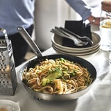 Eine Person reibt das Essen mit einer Reibe neben einer Pfanne mit gekochten Nudeln und Gemüse in einer Pfanne.