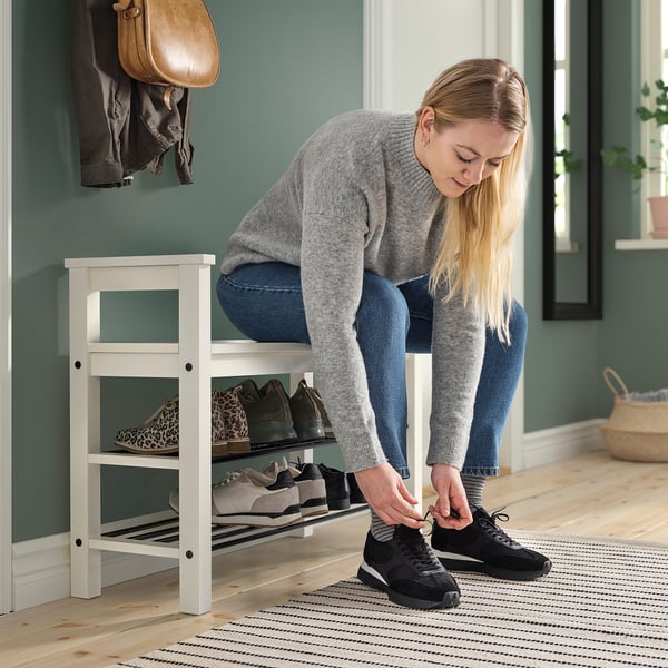 Frau bindet schwarze Turnschuhe, während sie auf weißer HEMNES-Bank in der Nähe des Schuhregals sitzt.