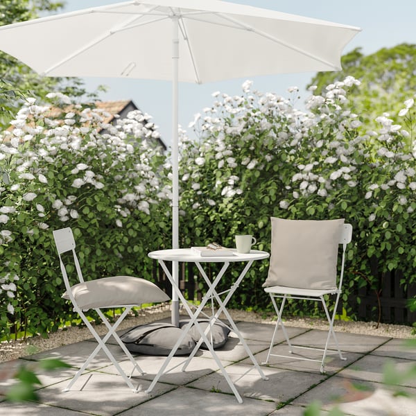 Sitzbereich im Freien mit weißen gepolsterten Stühlen und Sonnenschirm, umgeben von blühenden weißen Blumen.