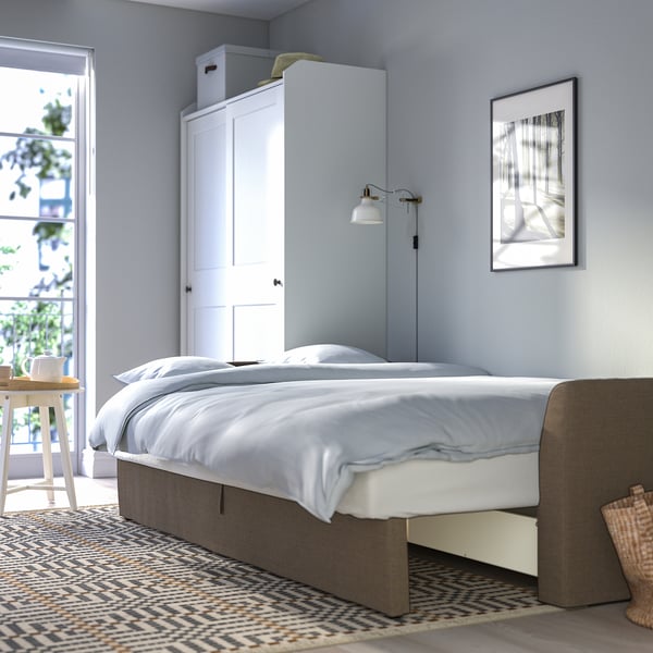 Schlafzimmer mit weißem Bett, grauem Bettrahmen, Wandlampe, hohem weißem Schrank, gemustertem Teppich und Wandkunst.