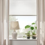Fenster mit lichtfilterndem Plissee, Lampe und Pflanzen auf Marmorbank.