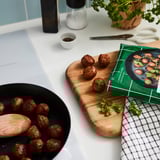 Eine Kochszene mit Fleischbällchen in einer schwarzen Pfanne und auf einem hölzernen Schneidebrett. Es gibt ein Rezeptbuch, Küchenutensilien und eine grüne Pflanze.