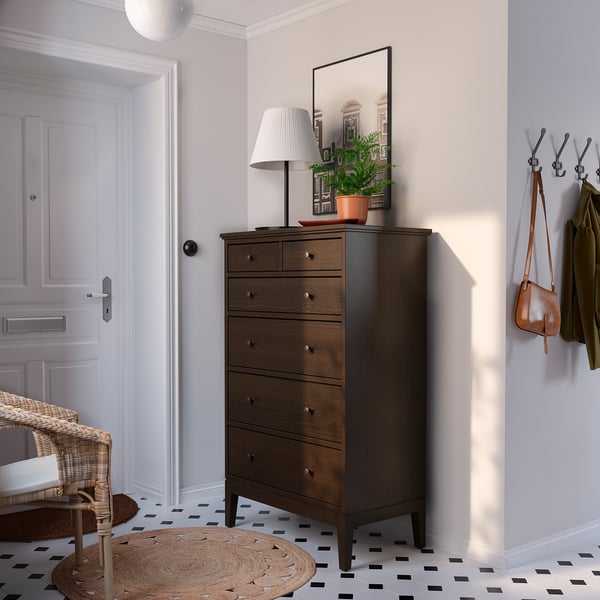 Moderner Eingangsbereich mit brauner IDANÄS-Truhe, weißer Lampe, grüner Pflanze, Kleiderhaken mit Jacken, schwarz-weißem Fliesenboden und Korbstuhl.