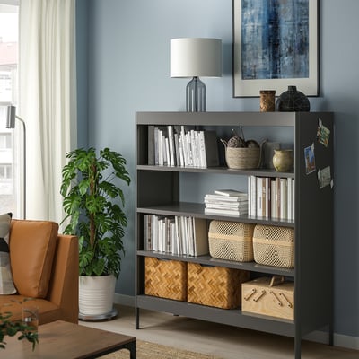 Schwarzes IDÅSEN Bücherregal mit verstellbaren Regalen an blauer Wand, in dem Bücher und Dekoration Platz finden, neben orangefarbenem Stuhl und Pflanze.