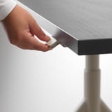 Eine Person stellt die Höhe eines IDÅSEN-Schreibtisches mit einem Griff ein und demonstriert so seine höhenverstellbare Funktion.