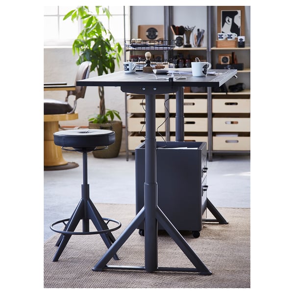 Ein moderner, höhenverstellbarer Schreibtisch mit grauer Platte, Metallbeinen und schwarzem Drehstuhl. Der Schreibtisch ist von Büromöbeln und Pflanzen umgeben.