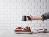 Eine Hand mit einem schwarzen IDEALISK-Sieb stäubt Puderzucker über einen mit Beeren belegten Kuchen an einer weiß gefliesten Wand.