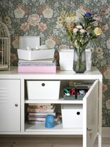 Weißer IKEA PS Metallschrank; florale Tapete; Platte: Aufbewahrungsboxen, Bücher, Vase mit Blumen; Unten: Offene Tür, die Bücher und Behälter freigibt.