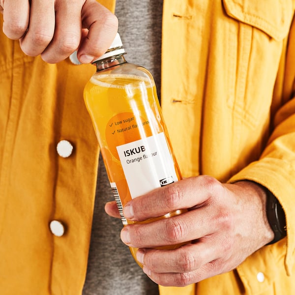 Person, die ein kohlensäurehaltiges Getränk mit Orangengeschmack öffnet. Die Trinkflasche ist mit ISKUB mit niedrigem Zuckergehalt und natürlichem Geschmack gekennzeichnet.