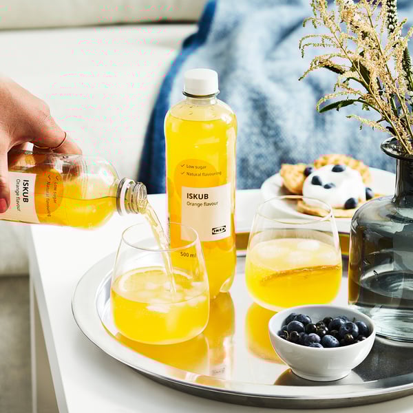 Die Hand gießt orangefarbenes ISKUB-Getränk in ein Glas, neben einem weiteren Glas, einer Flasche, Eis und einer Schüssel mit Blaubeeren auf einem Tablett.
