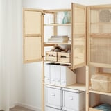 Ein Schrank aus Naturholz mit Bambustüren lässt sich öffnen und gibt den Blick auf Regale mit Aufbewahrungsboxen, Büchern und einer Karaffe frei.