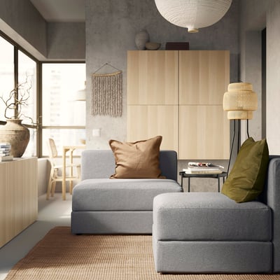 Modernes Wohnzimmer mit grauem modularem JÄTTEBO-Sofa, braunen Kissen, Holzschrank und Stehlampe. Minimalistisches Dekor mit natürlichem Licht.