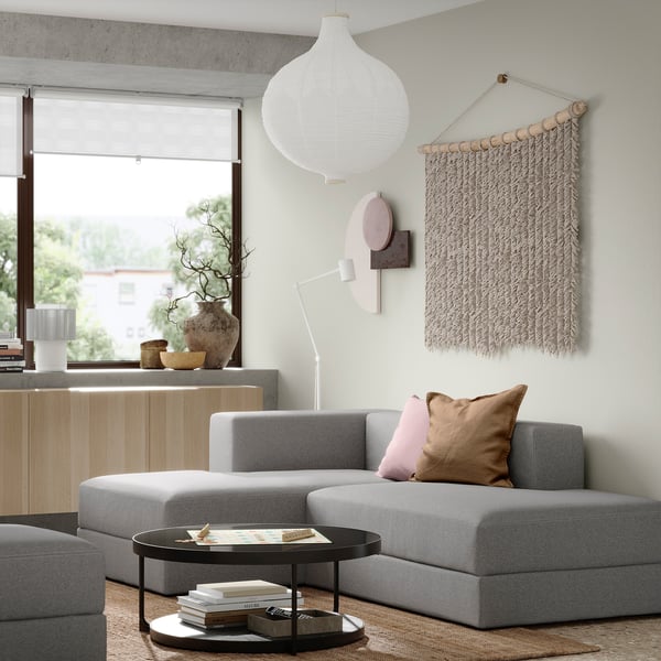 Modernes Wohnzimmer mit grauem JÄTTEBO-Sofa, rosa/braunen Kissen, schwarzem Couchtisch, Büchern, großem Fenster, Wandbehang.