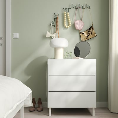 Modernes Schlafzimmer mit weißer Kommode, Lampe, Spiegel, Pflanzen und grünen Haken für Geldbörsen. Lichtvorhänge rahmen die Szenerie ein.