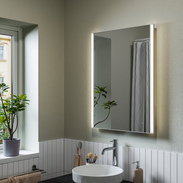 Der KÄRRSJÖN beleuchtete Spiegel hängt an einer hellgrünen Wand und spiegelt ein weißes Waschbecken und eine weiße Pflanze wider. Ihr sanftes Leuchten verstärkt die moderne Einrichtung.