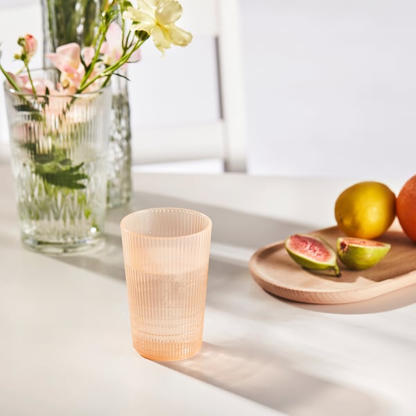 Ein pfirsichfarbenes Plastikglas steht auf einem Tisch neben einem Teller mit geschnittenen Früchten und einer Vase mit Blumen.