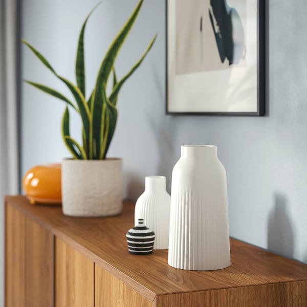 Holzschrank mit KAPKASTANJ-Vase, kleineren Vasen, Pflanze und Kunstwerk an hellblauer Wand.