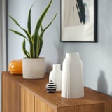 Holzschrank mit KAPKASTANJ-Vase, kleineren Vasen, Pflanze und Kunstwerk an hellblauer Wand.