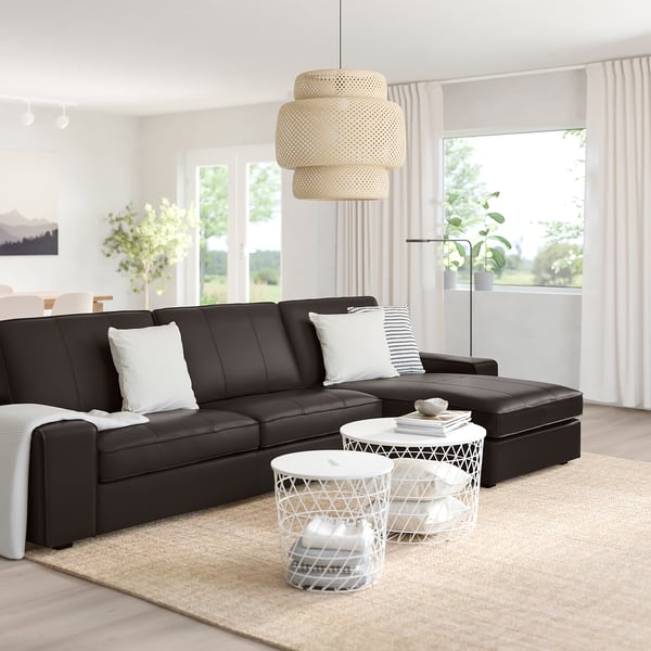 Modernes Wohnzimmer mit großem schwarzen KIVIK-Sofa, weißen Kissen, zwei Beistelltischen, großem Fenster, Lampe und Pflanzen.