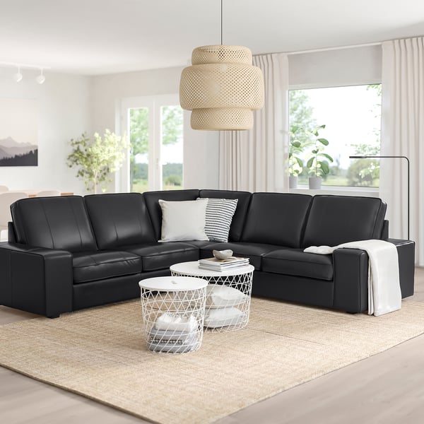 Modernes Wohnzimmer mit schwarzem L-förmigem KIVIK-Sofa, weißen Tischen, Teppich und Pendelleuchte. Große Fenster und Vorhänge. Gemütlicher Minimalismus.