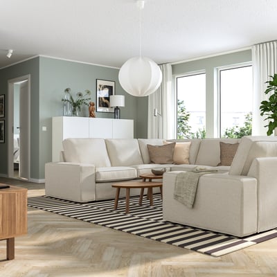 Modernes Wohnzimmer mit hellgrauem KIVIK-Sofa, gestreiftem Teppich, hölzernem Couchtisch, Pendelleuchte und großen Fenstern mit weißen Vorhängen.