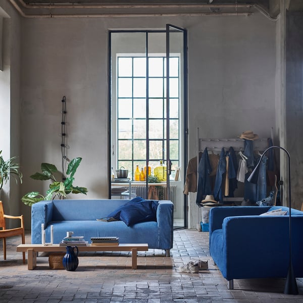 Modernes industrielles Wohnzimmer mit blauem KLIPPAN-Sofa und -Sessel, hölzernem Couchtisch, großem Fenster und Anlage.