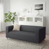 Modernes Wohnzimmer mit grauem KLIPPAN-Sofa, weißem Schrank, grüner Pflanze und Teppich.