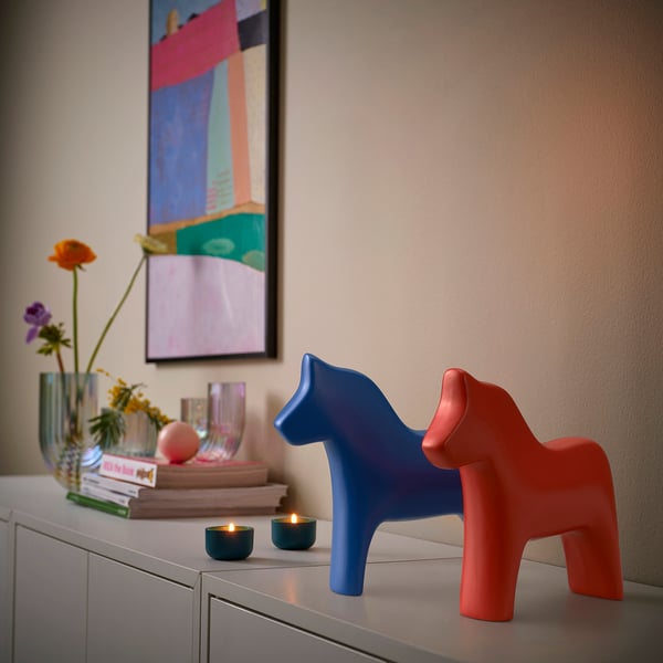 Zwei Dala-Pferde aus Keramik, eines blau, eines rot, auf einem Sideboard mit Büchern und Blumen.