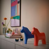 Zwei Dala-Pferde aus Keramik, eines blau, eines rot, auf einem Sideboard mit Büchern und Blumen.