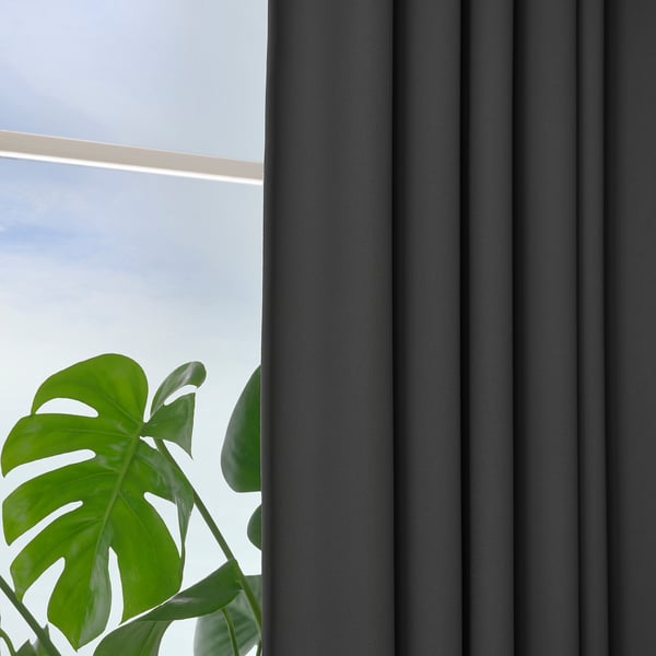 Dunkelgraue Vorhänge mit geflochtener Textur bedecken teilweise ein Fenster, so dass etwas blauer Himmel zu sehen ist. In der unteren linken Ecke ist eine grüne Pflanze zu sehen.