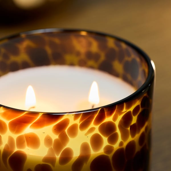 Eine entzündete Kerze in einem gemusterten Glasbehälter, die mit drei Dochten ein warmes, einladendes Licht ausstrahlt und hochwertige Handwerkskunst zeigt.