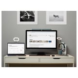 Ein eleganter weißer Schreibtisch besteht aus einem Monitor, einem Laptop und einer LED-Lampe. Dahinter hängen zwei gerahmte Schwarz-Weiß-Fotos.