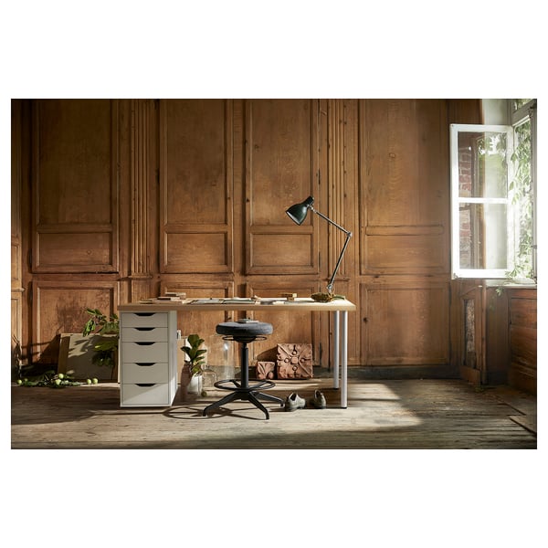 Kompaktes Home-Office-Setup: weißer LAGKAPTEN-Schreibtisch mit Schubladenschrank, schwarzem Drehstuhl und verstellbarer Lampe an einer Holzpaneelwand mit Fenster.