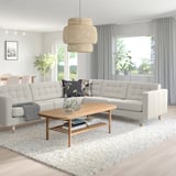 Modernes Wohnzimmer mit hellgrauem L-förmigem Sofa, Couchtisch aus Holz und Pendelleuchte. Graue Vorhänge rahmen die Fenster ein und lassen natürliches Licht herein.