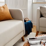Gemütliches Wohnzimmer mit beigem Sofa, gelbem Kissen, blauem Überwurf und Holzkorb auf Teppich am Fenster.