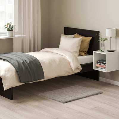 Schlafzimmer mit LINDKNUD beigem Teppich, weißem Bett, schwarzem Kopfteil und Nachttisch mit Lampe. Der Teppich ist klein und passt unter das Bett.