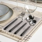 Moderner Tisch mit LÖFTESRIK Besteck auf beigem Tischset. Inklusive Gabel, Messer, Löffel und leerem Weinglas.