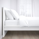 Minimalistisches weißes Bett mit Kopfteil, zwei Kissen und gefalteter Decke auf Holzboden.