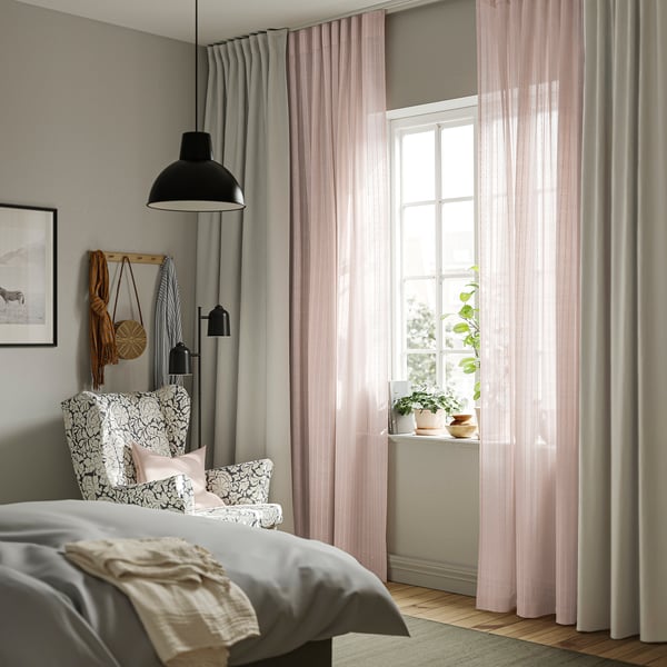 Schlafzimmer mit MATILDA-transparenten rosa Vorhängen, gestreift und sprudelnd, neben einem großen Bett mit gemusterten Kissen, Wandlampe und nahegelegenen Pflanzen.