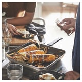 Eine Person, die gegrillten Mais in einer MIDDAGSMAT-Pfanne würzt, die auf einem Esstisch mit Tellern und Gläsern steht.