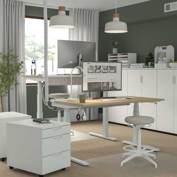 Moderner Sitz-Steh-Schreibtisch mit zwei Monitoren, Pendelleuchten und minimalistischem Dekor. Ergonomischer Hocker. Heller, sauberer Arbeitsplatz.