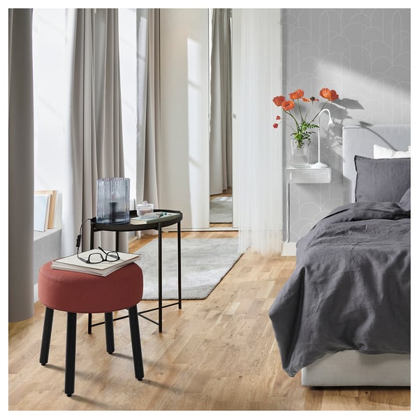 Schlafzimmer mit rotem MÖRTFORS-Hocker, grauem Bett, schwarzem Beistelltisch, Vase mit Blumen und großem Fenster.
