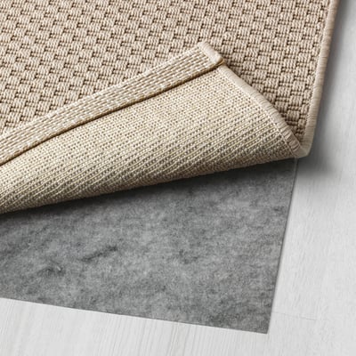 Ein beiger gewebter Teppich mit einer gefalteten Ecke, der über eine graue Filzunterlage gelegt wird und für den Innen- und Außenbereich geeignet ist.