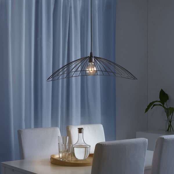 Modernes Esszimmer mit MOTSJÖ-Lampe, Drahtschirm, klarer Glühbirne, weißen Stühlen, blauen Vorhängen und Holztablett mit Karaffe und Gläsern.