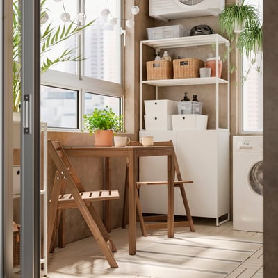 Moderne Küchenecke mit NÄMMARÖ Tisch, Stühlen, Tassen, Pflanzen und Regalen für Körbe und Reinigungsmittel. Weiße Geräte, Fliesenboden.