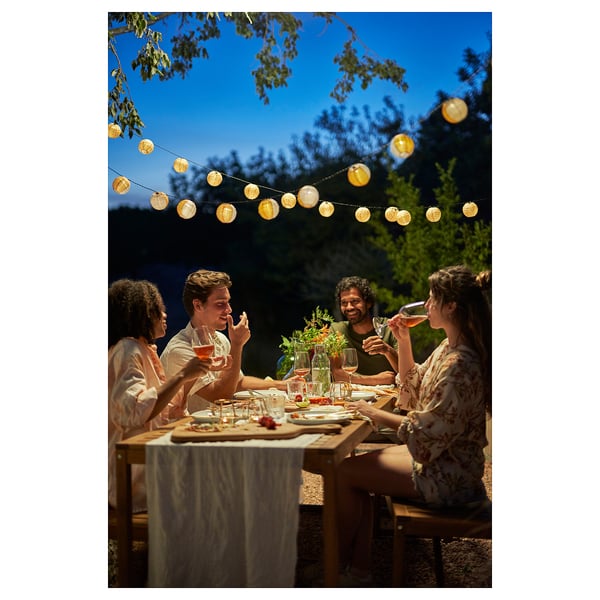 Vier Freunde stoßen unter Papierlaternen an einem Tisch im Freien an, der zum Essen gedeckt ist.