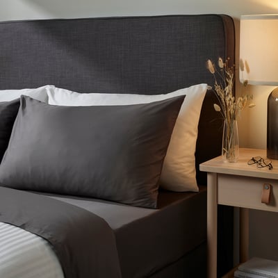 Ein modernes Schlafzimmer mit einem dunkelgrauen Kopfteil und Bettwäsche sowie einem Nachttisch aus Holz mit getrockneten Blumendekorationen und einer Lampe.