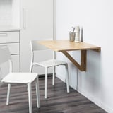 Klappbarer Küchentisch aus Holz mit zwei weißen Kunststoffstühlen vor einer weißen Wand.