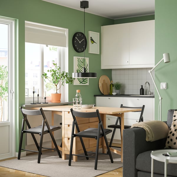 Moderne Küche mit Tageslicht, Holztisch, schwarzen Stühlen und weißen Schränken. Grüne Wand mit Uhr, Kunst und Pflanze. Graue Couch in der Nähe.